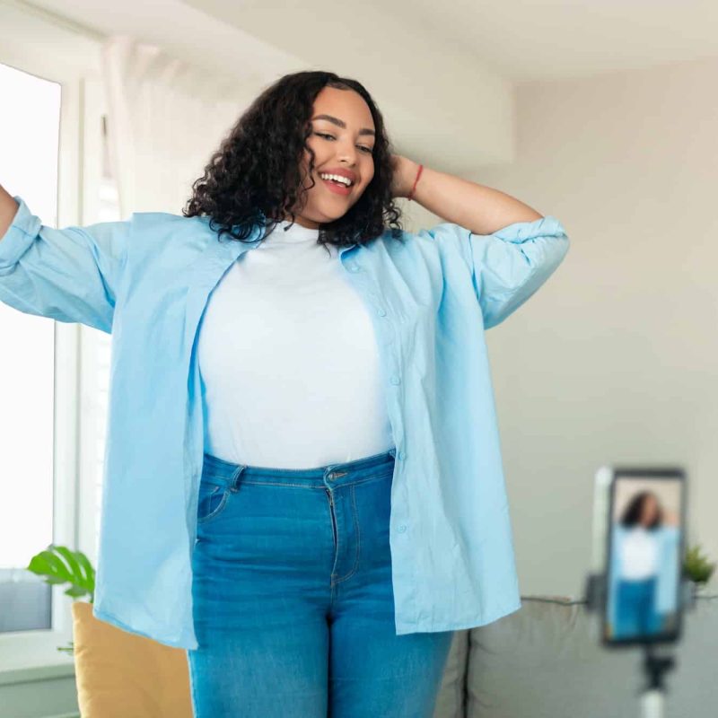 African american plus size female influencer recording video on smartphone, dancing on camera at