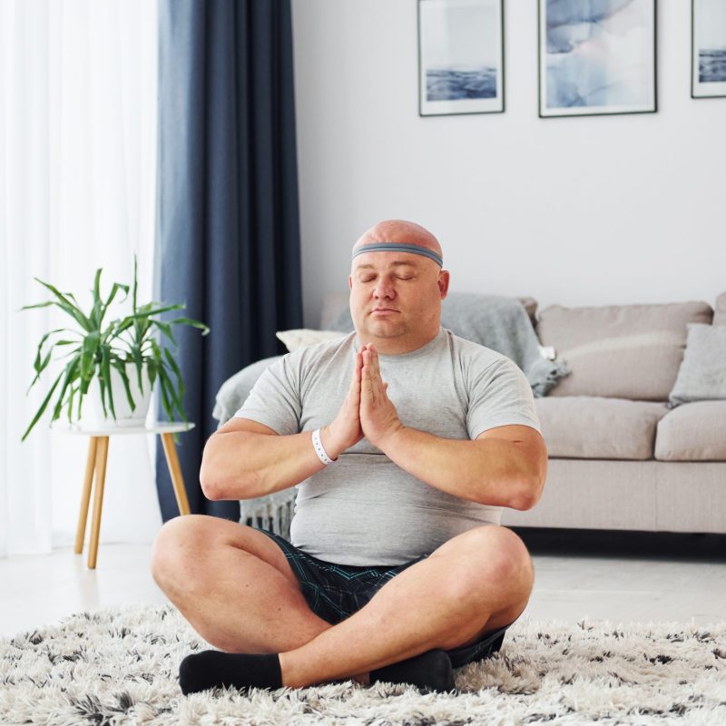 Doing yoga exercises. Funny overweight man in casual clothes is indoors at home.