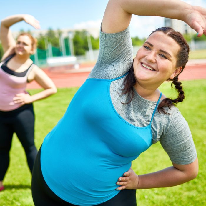 Happy young chubby women doing side-bends on green lawn on sunny day