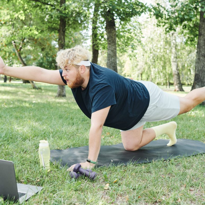 Overweight man exercising with dumbbells on exercise mat in the park and watching sports training online on laptop